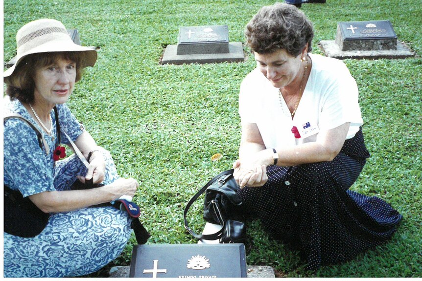 Two women, one wearing a hat, sit next to a grave. One looks at the camera. Other graves behind.