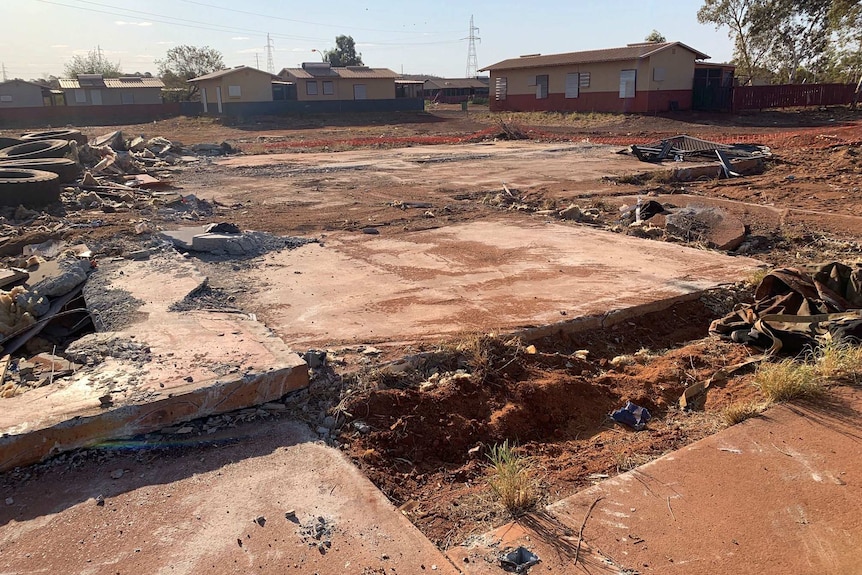 A bulldozed block of land with rubble, tyres, red dirt, and the foundations of buildings, with houses in the background.