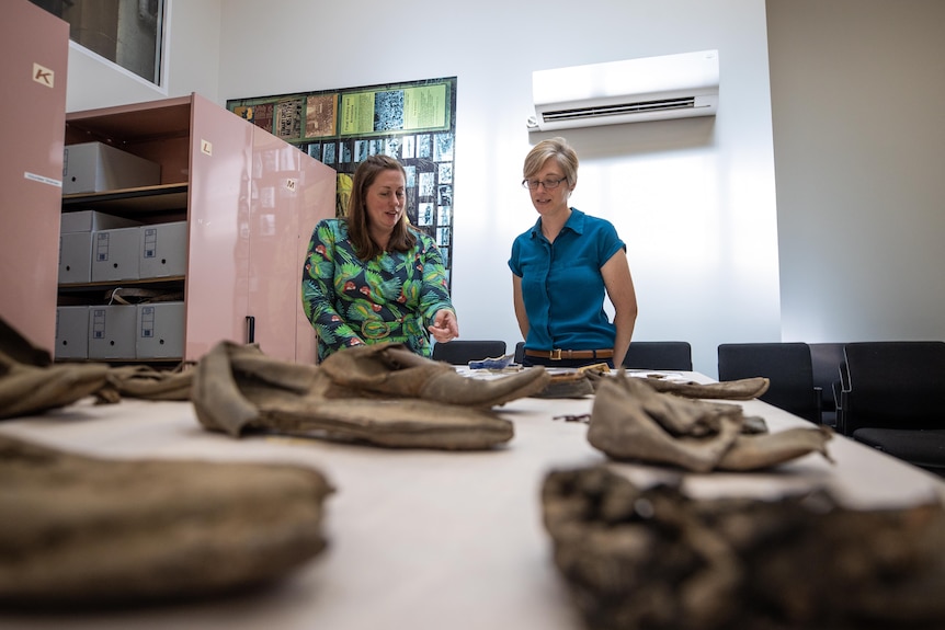 Two women stand at the end of a table, talking. A range of old, leather shoes are arrayed on the table in front of them.