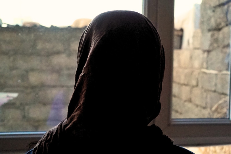 A woman in Islamic headscarf stands at a window with her back to the camera