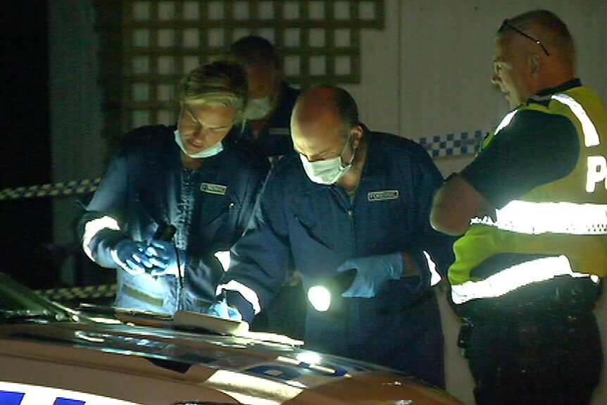 Three police officers gather around the bonnet of a police car and shine a torch on some documents.
