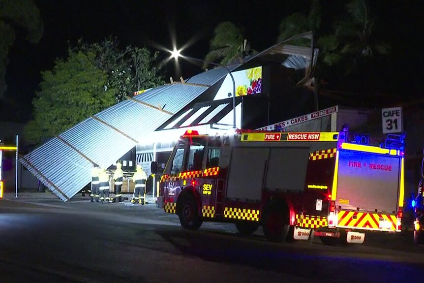 Fire trucks and firefighters on the scene of a shop with a large tin roof hanging off its edge