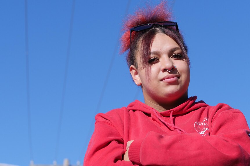 A teen in a red hoodie poses under a blue sky with her arms crocce