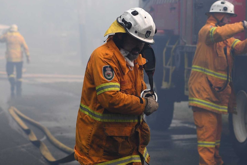 A firefighter pulls a hose, a firetruck and smoke are in the background.