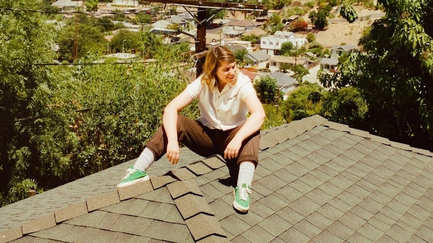 Singer Alex Lahey is wearing brown pants, a cream button-up top and green sneakers as she sits on a brown tiled rooftop.