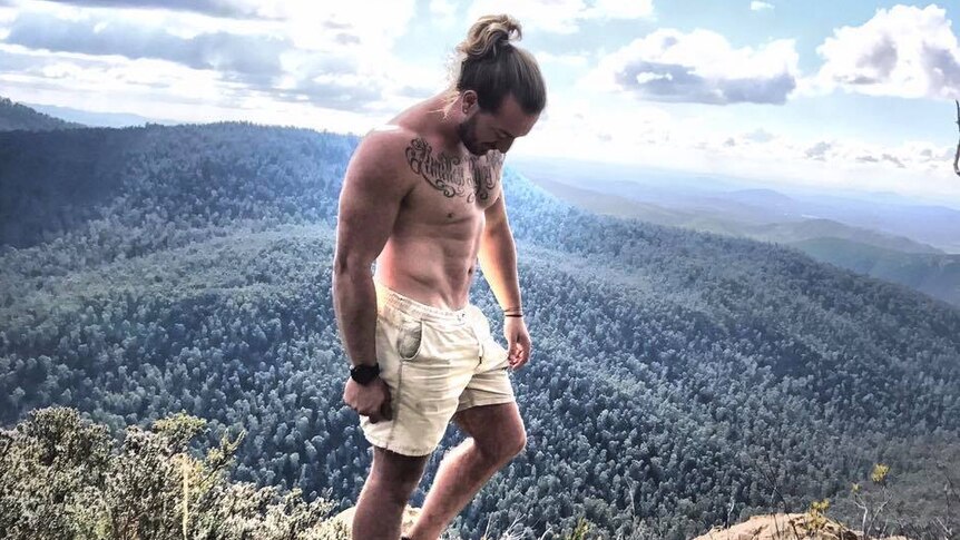 A man stands shirtless on the top of a mountain. He has a large written tattoo across his chest.