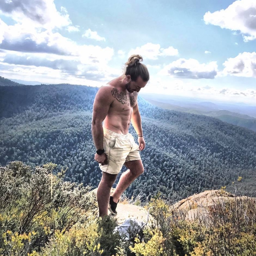 A man stands shirtless on the top of a mountain. He has a large written tattoo across his chest.