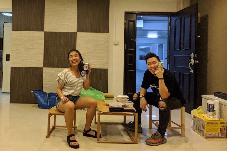 Ken Yip and his wife sit on small stools drinking beer and smiling at the camera in their new empty apartment.