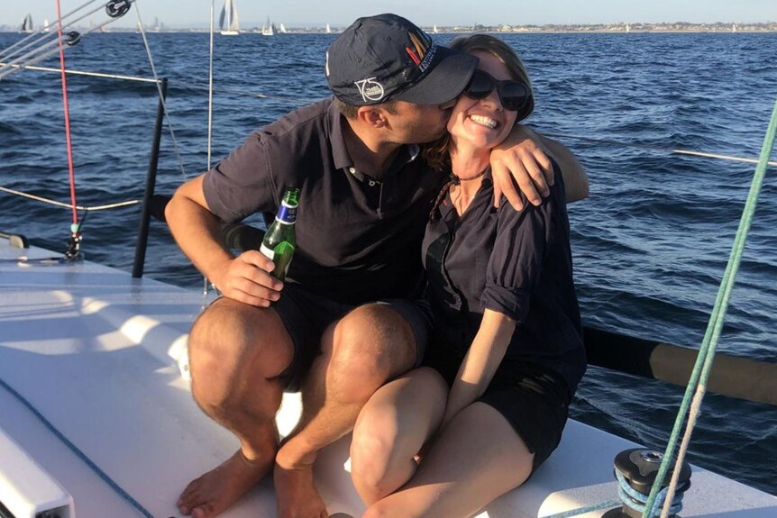 Cameron Dale with his arm around smiling Jessica Watson, sitting on a sailboat, water behind.