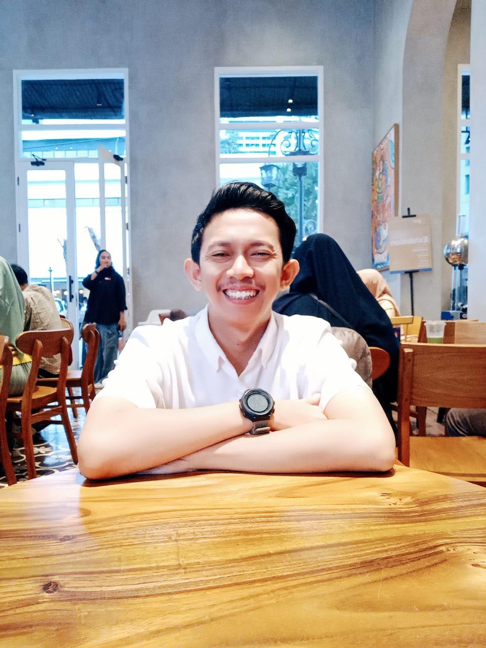 A young man wearing white shirt, sitting in the middle of a cafe, looking at the camera, smiling.