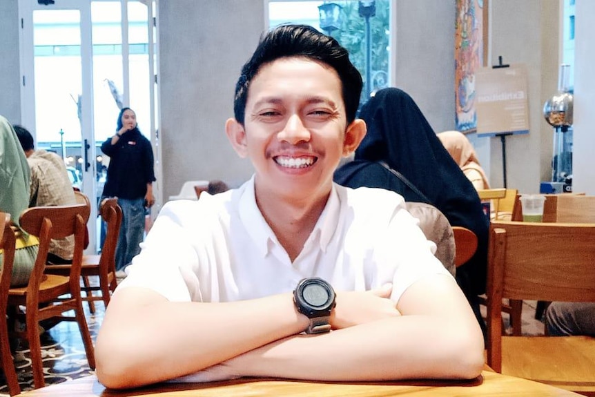 A young man wearing white shirt, sitting in the middle of a cafe, looking at the camera, smiling.