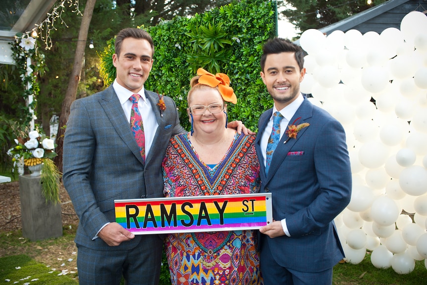 Two men hold a 'Ramsay St' sign and smile happily as Magda Szubanski stands between them.