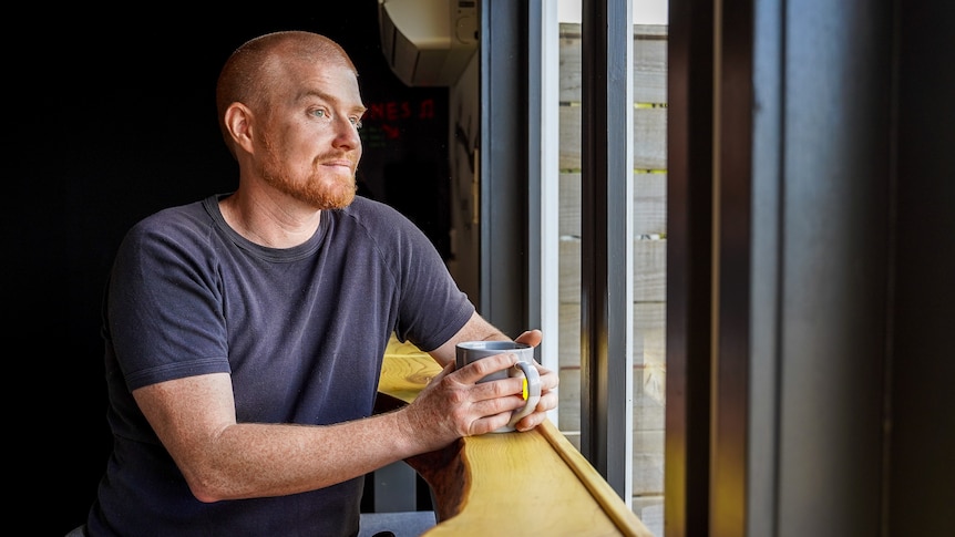 A man sits at a bench looking out of a window, coffee cup in his hands.