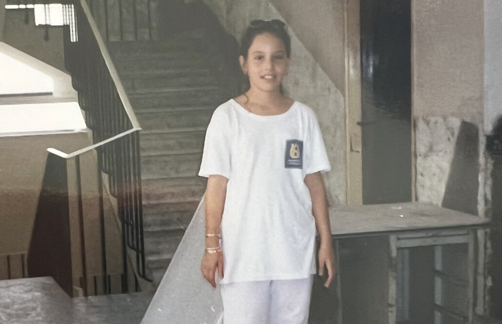 Girl with brown hair tied back stands near stairwell wearing white T-shirt.