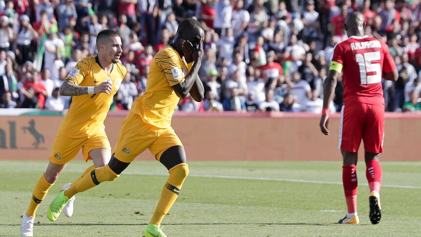 Awer Mabil covers his mouth with his hand and taps his forehead as Jamie Maclaren follows him after scoring for the Socceroos