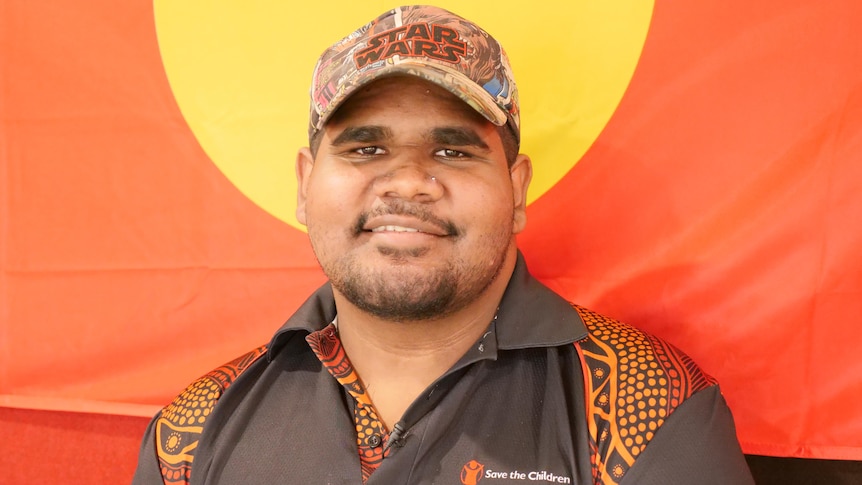 An Aboriginal man wearing a Star Wars cap and Indigenous patterned shirt, standing in front of an Aboriginal flag