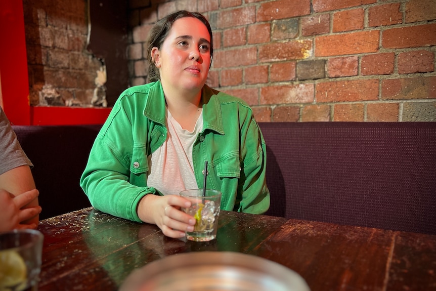 A woman wearing a bright green jacket and white T-shirt, with her dark hair pulled back, sitting at a pub with a drink