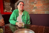 A woman wearing a bright green jacket and white T-shirt, with her dark hair pulled back, sitting at a pub with a drink
