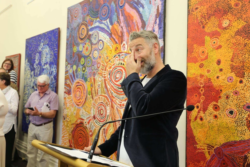 Middle-aged man with white and grey beard wipes a tear from his face from behind a lectern.