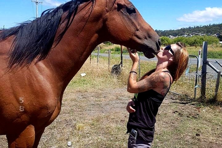 A woman with long brown hair kisses the nose of a horse in a paddock