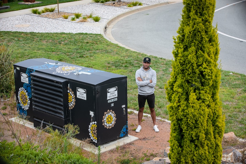 A man wearing a black cap standing beside a substation that has Indigenous artworks painted on it. 