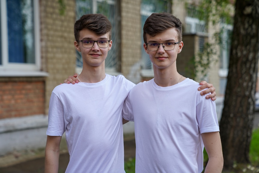 Two teenage boys, both wearing white shirts, glasses and sporting identical brown haircuts, smile at the camera