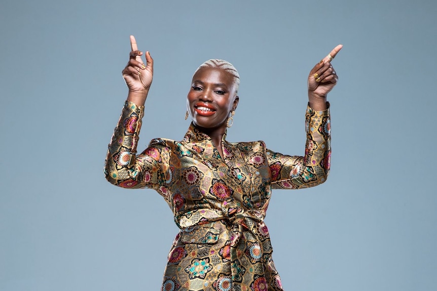 A woman in a gold dress and white hair is smiling and pointing up, like she's dancing.