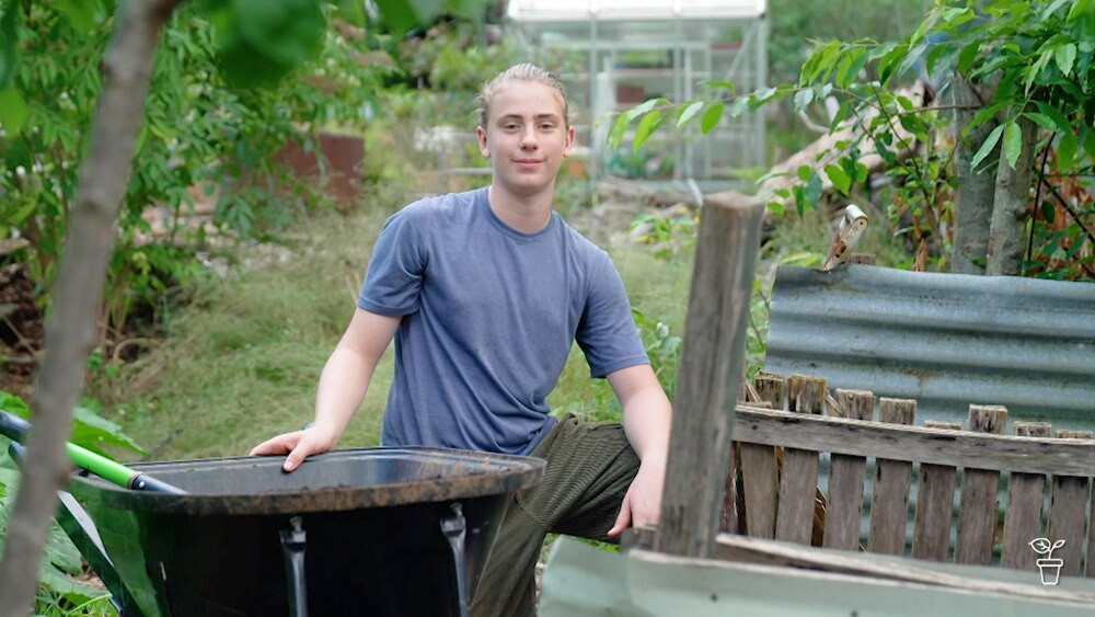 A youg gardener sitting in the garden next to a wheelbarrow and  a compost bay.
