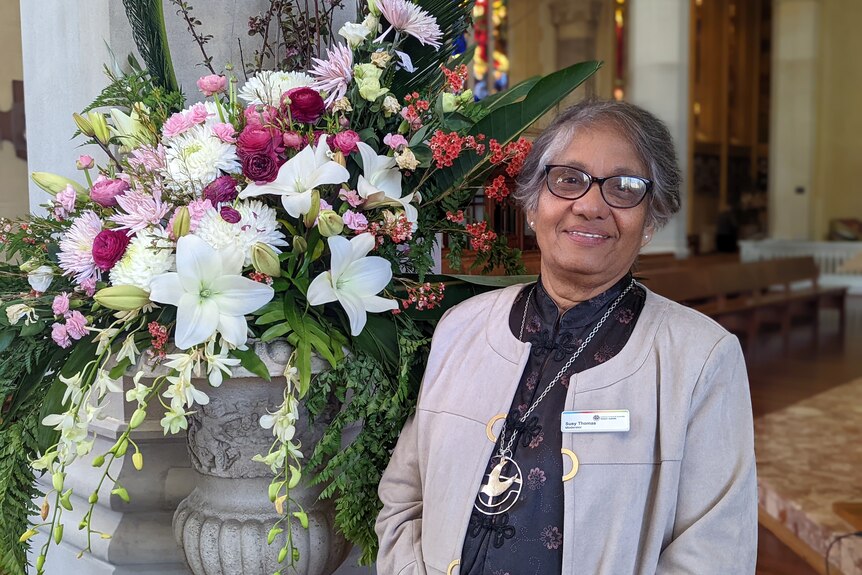 Smiling south Asian woman wears light jacket, dark top with gold print, stands next to floral arrangement, wooden pews behind.