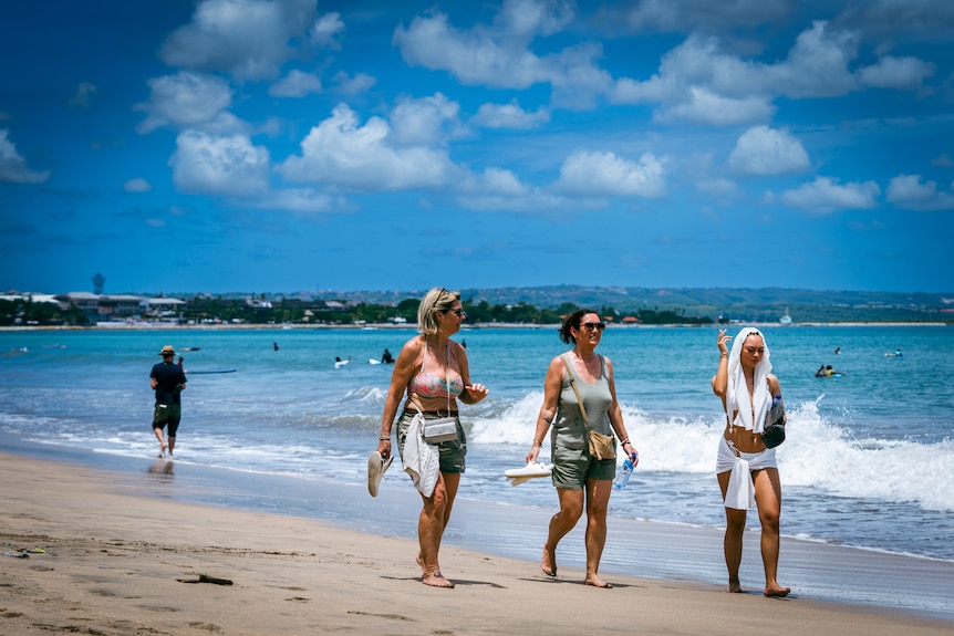 Three girls walk along a white sandy beach on a sunny day.