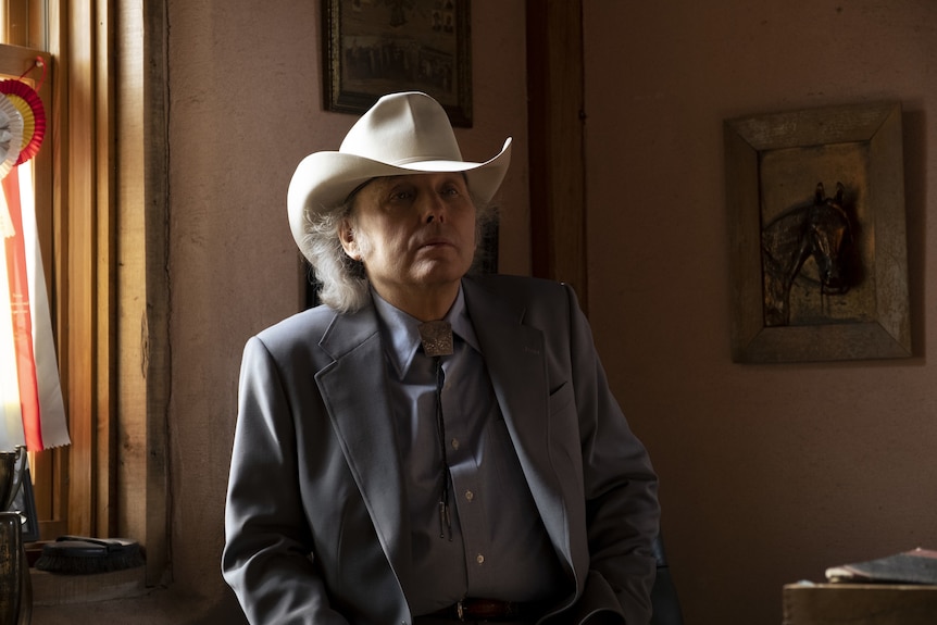 A 60-something man in a cowboy hat and suit stands looking nonplussed in an office space decorated with a painting of a horse