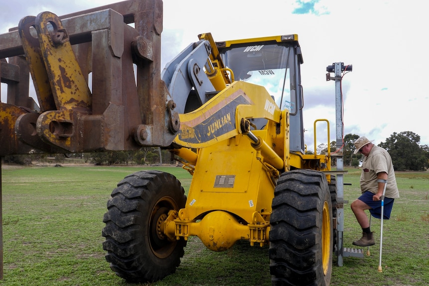 A farmer who only has a left leg using a hoist to climb into the cab of his tractor.