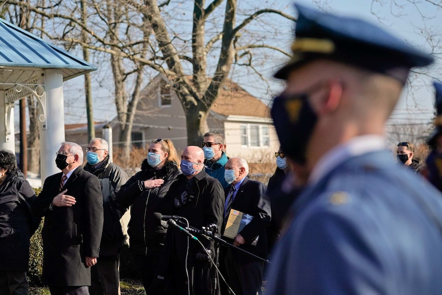 A man wearing a police uniform looks on as a group of people in black wearing masks hold their hands to their hearts