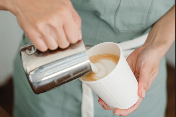 Manos de mujer vertiendo café de una jarra plateada en una taza de café blanca para llevar