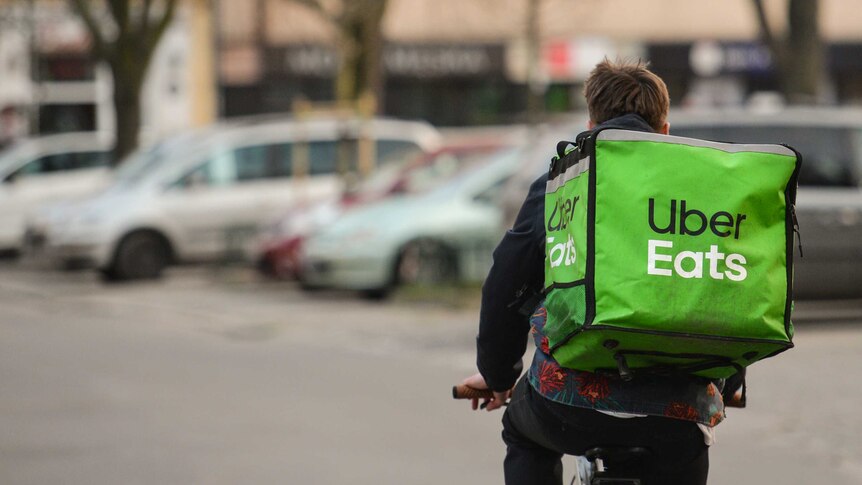 A young male Uber Eats delivery driver is seen from behind as he rides a bicycle at an intersection