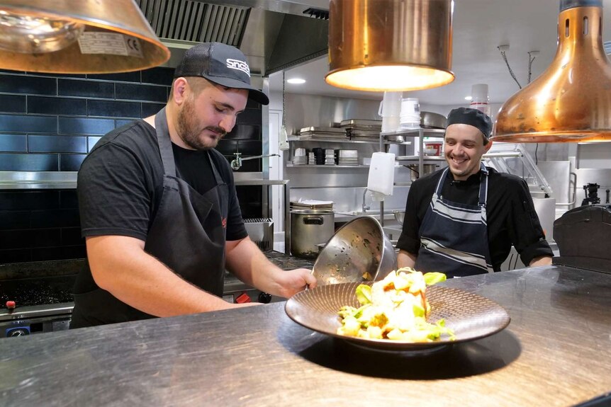 Two chefs wearing black t-shirts and aprons serve up a dish in a restaurant kitchen