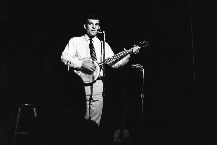 Steve Martin holds a banjo on stage in front of a mic wearing light colours, surrounded by an audience shrouded in darkness.