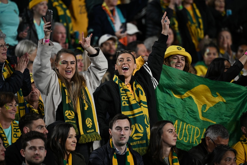 Two women wearing green and gold scarves stand and cheer in the grandstands.
