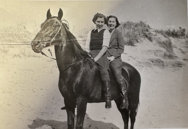 Two women ride a black horse on the beach in a black and white photo