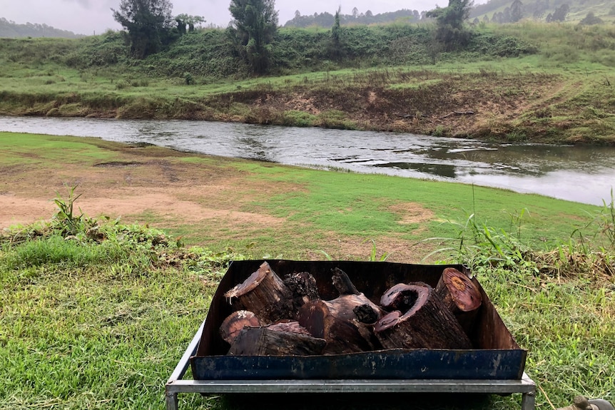 Fire pit with logs in it in the foreground with river and riverbank in background.