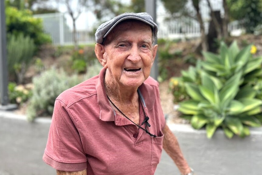 An elderly man wearing a pink polo shirt and a grey hat smiles for the camera in front of a garden bed.