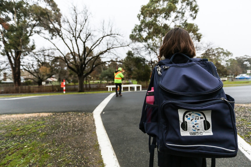 A primary schoolgirl stands facing the road. Her backpack is in foreground. School crossing supervisor is ahead of her.