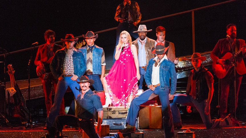 Kylie Minogue sings on stage, flanked by seven men dressed as cowboys