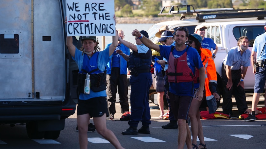 Kayakers with lifejackets walk on strip of road holding sign saying 'we are not the criminals'