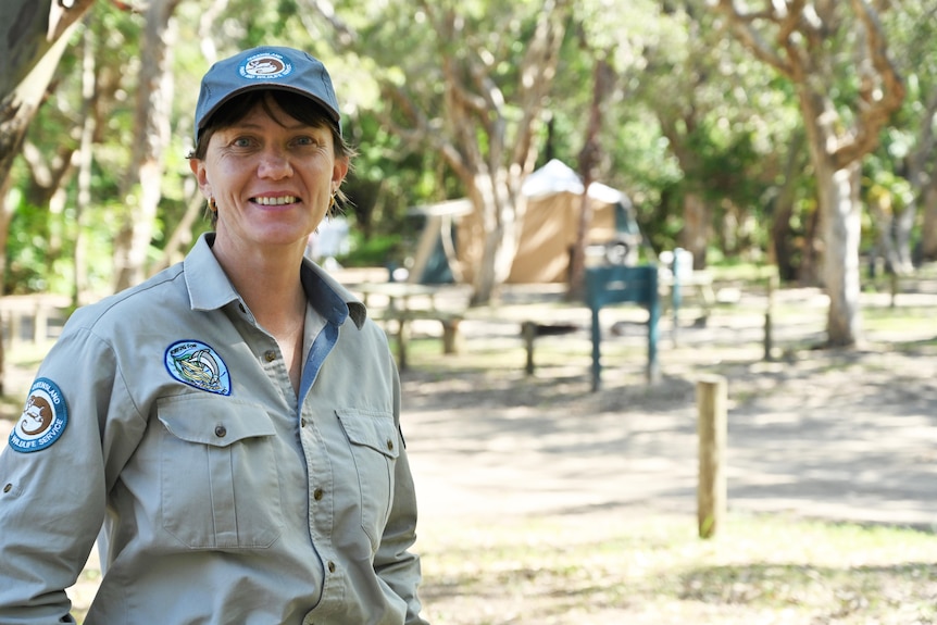 A woman in khaki ranger uniform and dark green cap smiling while standing in a treed campground