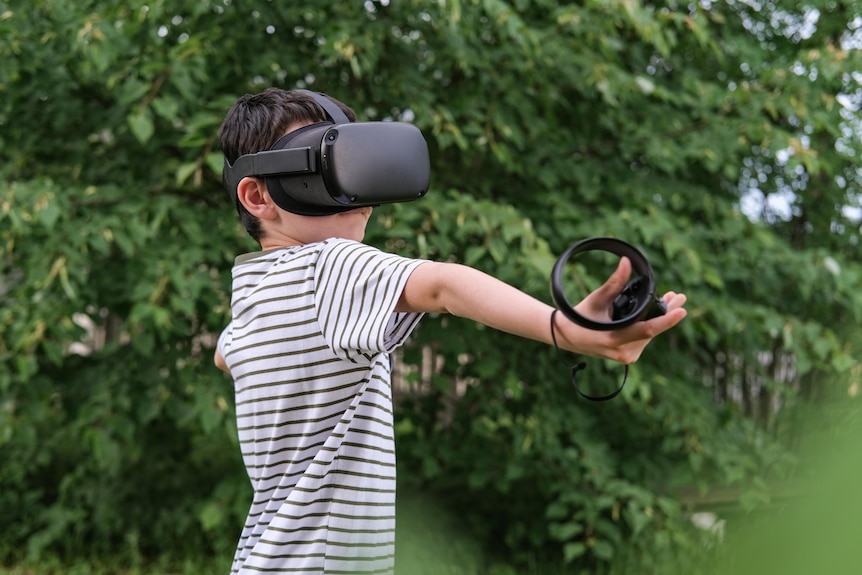 A seven-year-old boy is outside and wearing a virtual reality headset. He outstretches an arm holding a VR controller.