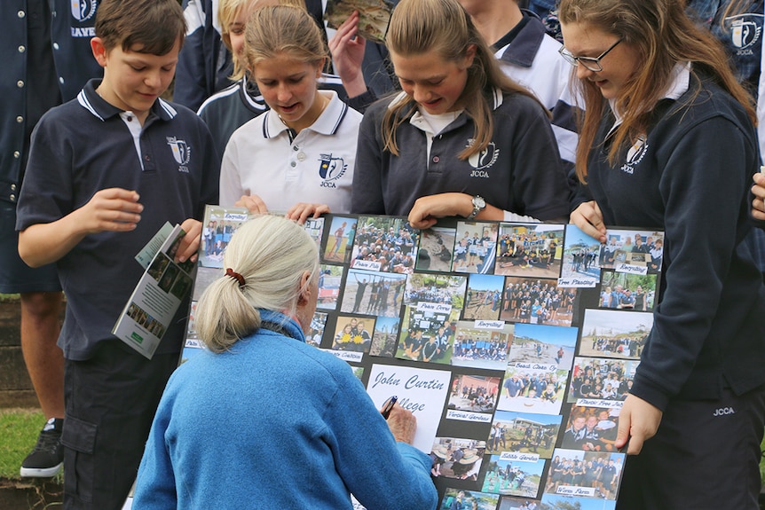 Four school children show a photo board to Dr Jane Goodall, who signs it for them.