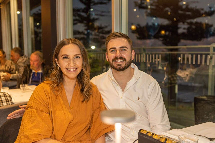 Michelle in an orange top, Jono in a white shirt sitting next to each other at a table, smiling.