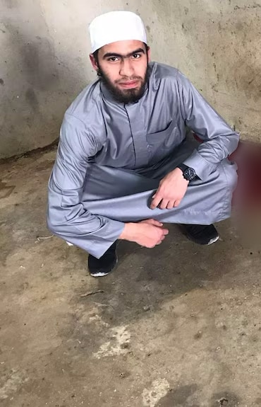 A man wearing a traditional Muslim cap and long shirt crouches on a concrete floor. He is looking at the camera.