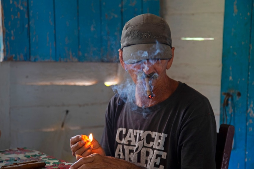 And older tobacco farmer lights a cigar on his farm in Vinales, Cuba. Smoke rises in front of his face as he does this.